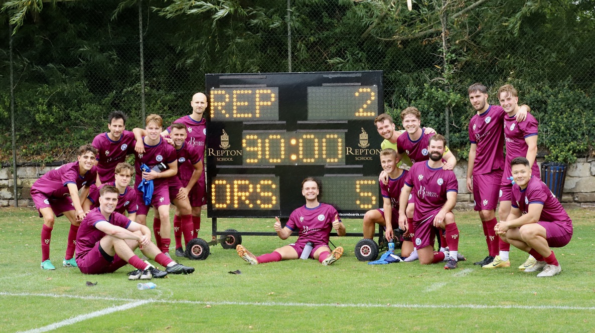 Eine Fußballmannschaft in kastanienbraunen Trikots posiert nach einem Spiel auf einer Wiese mit Bäumen und einem Zaun im Hintergrund fröhlich vor einer Anzeigetafel, auf der REP 2 ORS 5 steht.