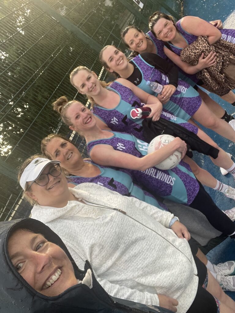 Un gruppo di sette donne, alcune delle quali indossano abiti da netball personalizzati dei Whirlwinds nei toni del blu e del viola, sorridono insieme all’aperto su un campo sportivo bagnato; in primo piano, una donna con una giacca con cappuccio sta scattando un allegro selfie.