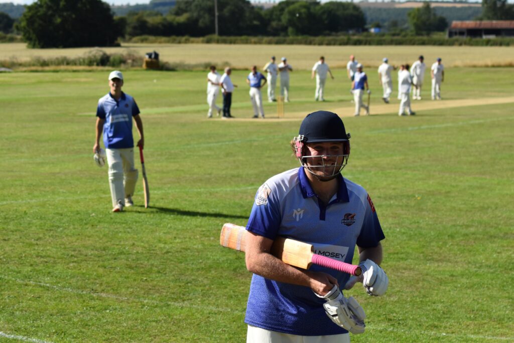 Ein Cricketspieler in blau-weißer Spielkleidung, der einen Helm trägt und einen Schläger in der Hand hält, verlässt lächelnd das Spielfeld. Im Hintergrund sind weitere Spieler auf einem grünen Cricketfeld unter strahlend blauem Himmel zu sehen.