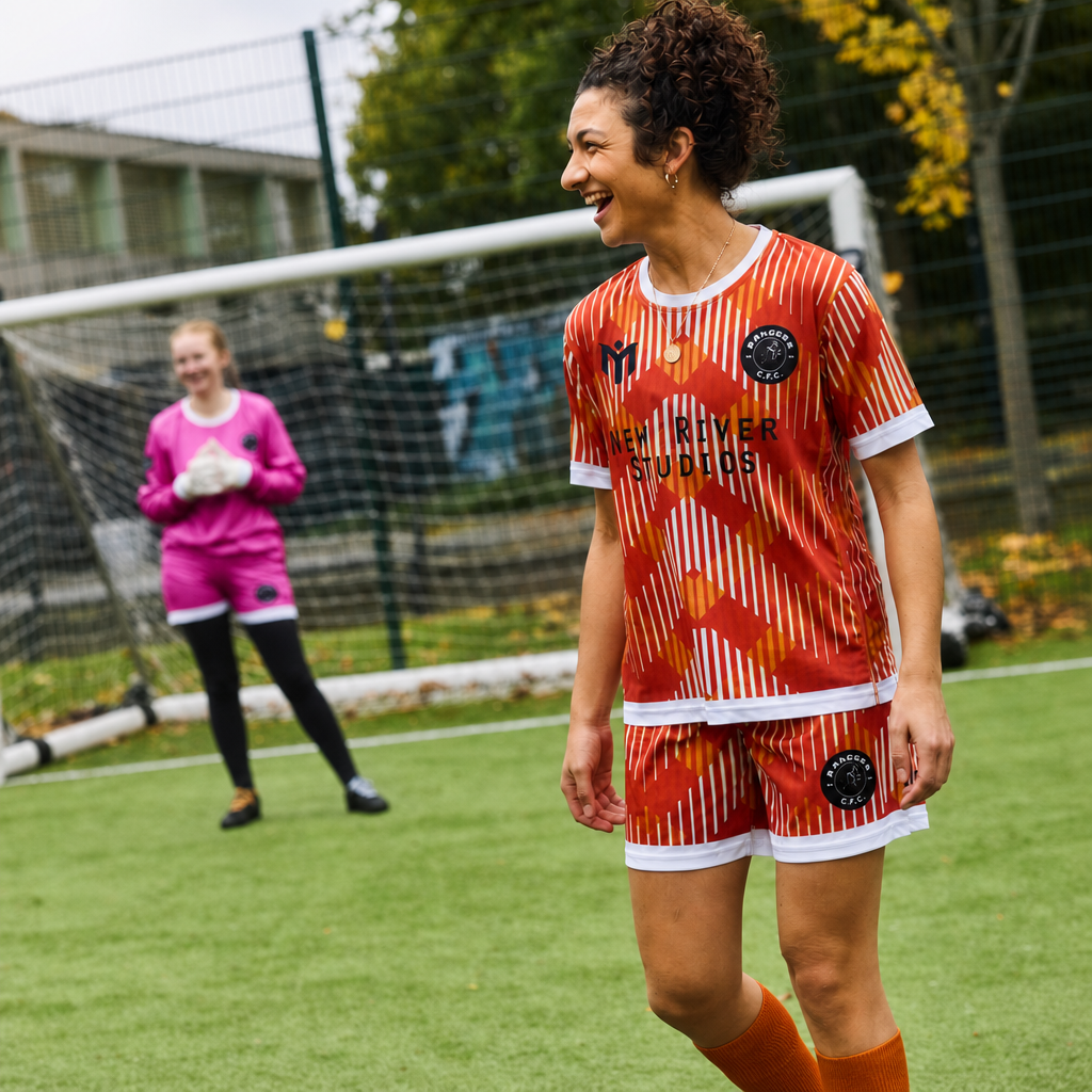 A smiling woman in an orange and white football kit stands on a football pitch, with a goalkeeper in pink and black kit near the goal in the background. Trees and fencing are visible behind them.