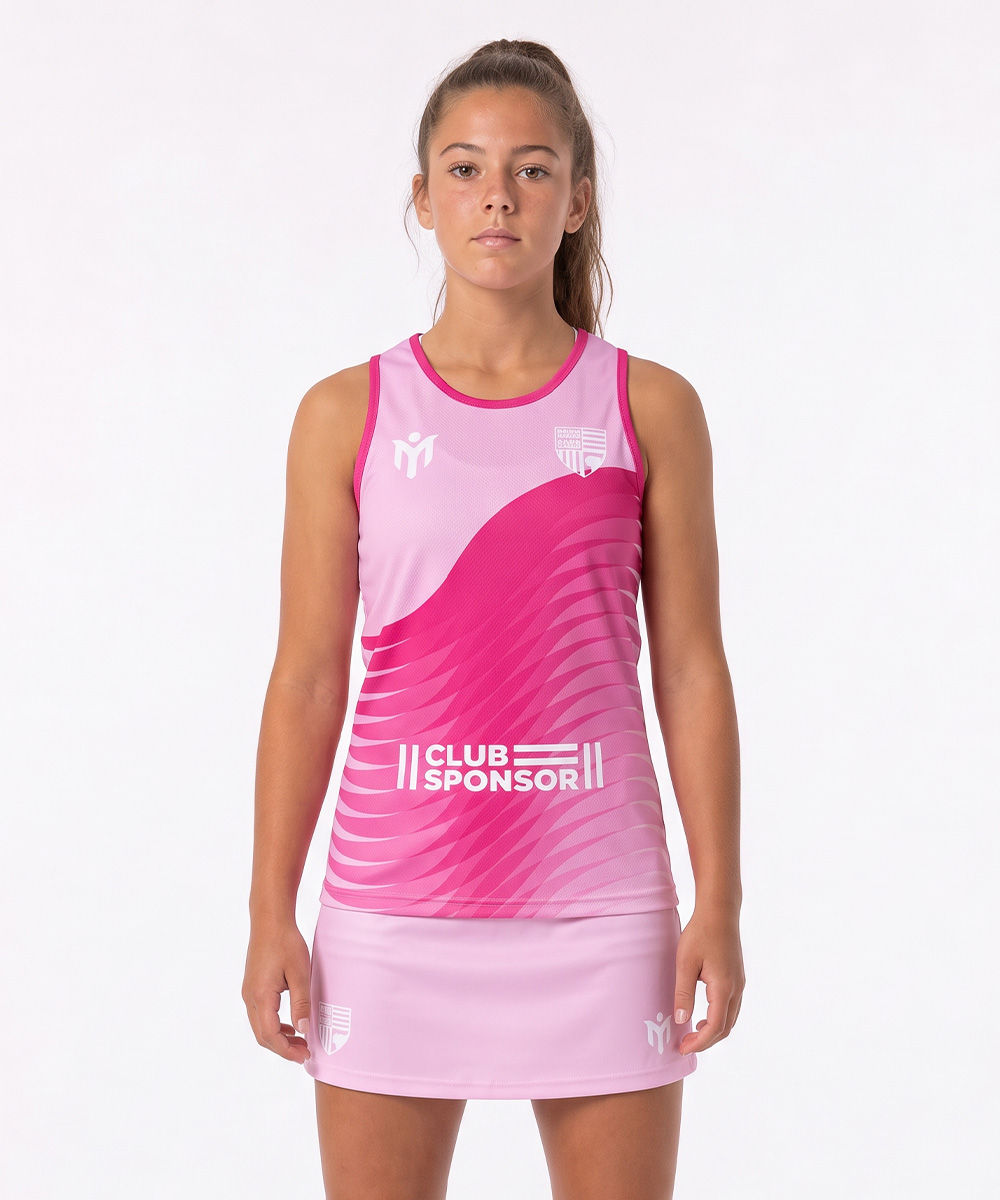 Una giovane donna è in piedi, rivolta in avanti, e indossa un abito sportivo rosa con motivi bianchi e la scritta "CLUB SPONSOR" sul davanti; indossa eleganti canotte da netball su uno sfondo bianco a tinta unita.