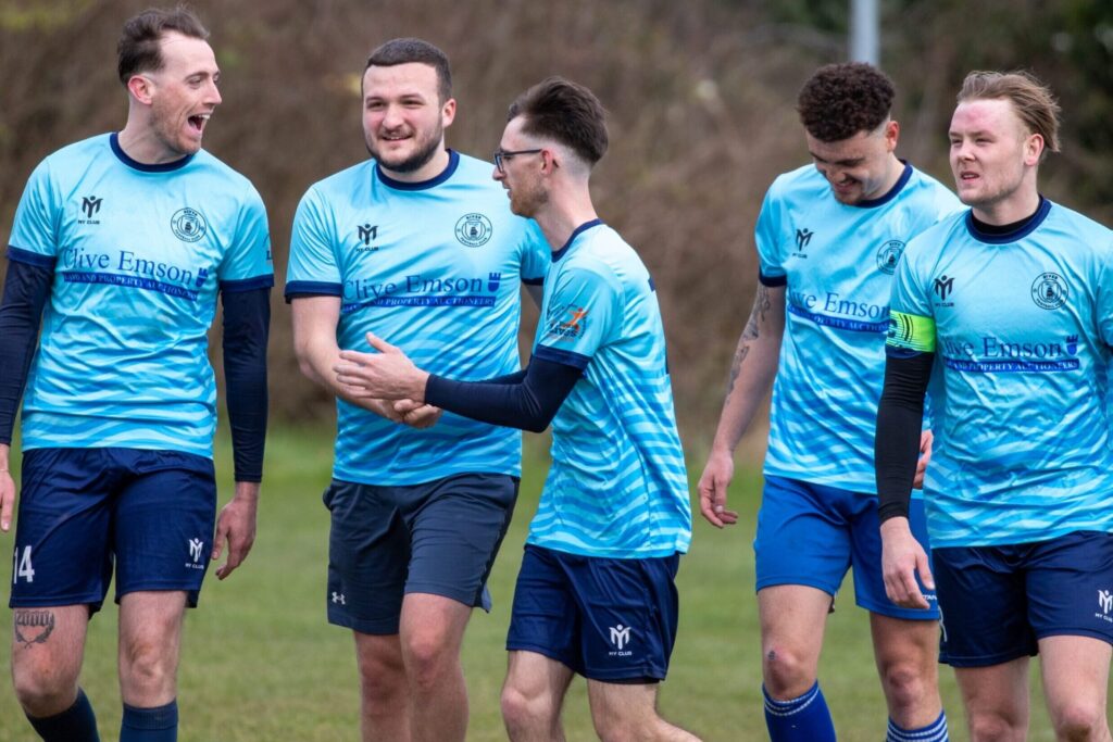 Eine Gruppe männlicher Fußballspieler in hellblauen Trikots läuft gemeinsam über einen Rasenplatz, lächelt und unterhält sich, während ein weiterer Spieler in einem grünen Trikot in der Nähe entlangläuft – ein Bild, das die Stimmung der bevorstehenden AI-FIFA-Weltmeisterschaft 2026 einfängt. Im Hintergrund sind Bäume und Sträucher zu sehen.