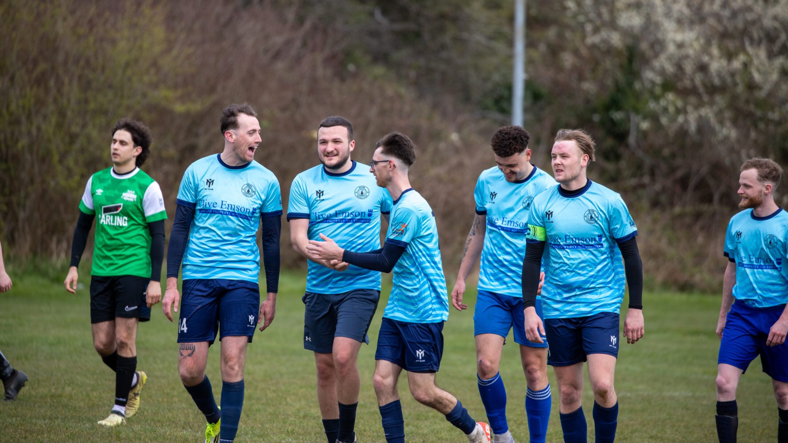 Un gruppo di calciatori in maglia azzurra cammina insieme su un campo in erba, sorridendo e chiacchierando, mentre un altro giocatore in maglia verde cammina lì vicino, incarnando lo spirito della prossima Coppa del Mondo FIFA 2026 con intelligenza artificiale. Sullo sfondo si vedono alberi e cespugli.