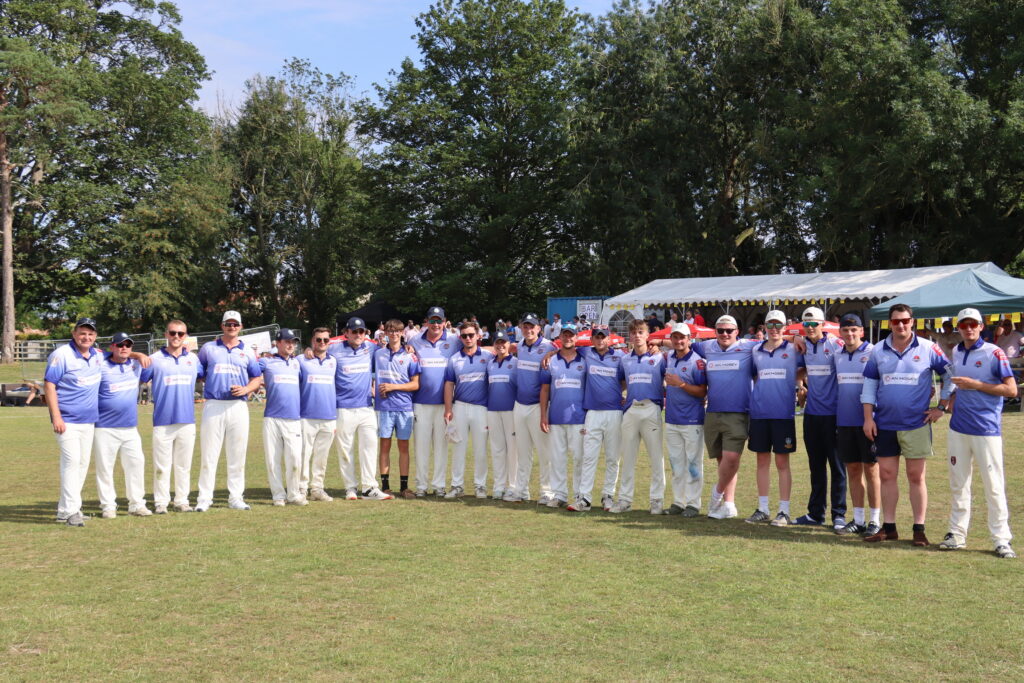 Eine große Gruppe von Männern in einheitlichen blau-weißen Cricket-Trikots posiert gemeinsam auf einer Wiese, im Hintergrund sind Bäume und ein weißes Festzelt zu sehen – an einem sonnigen Tag.