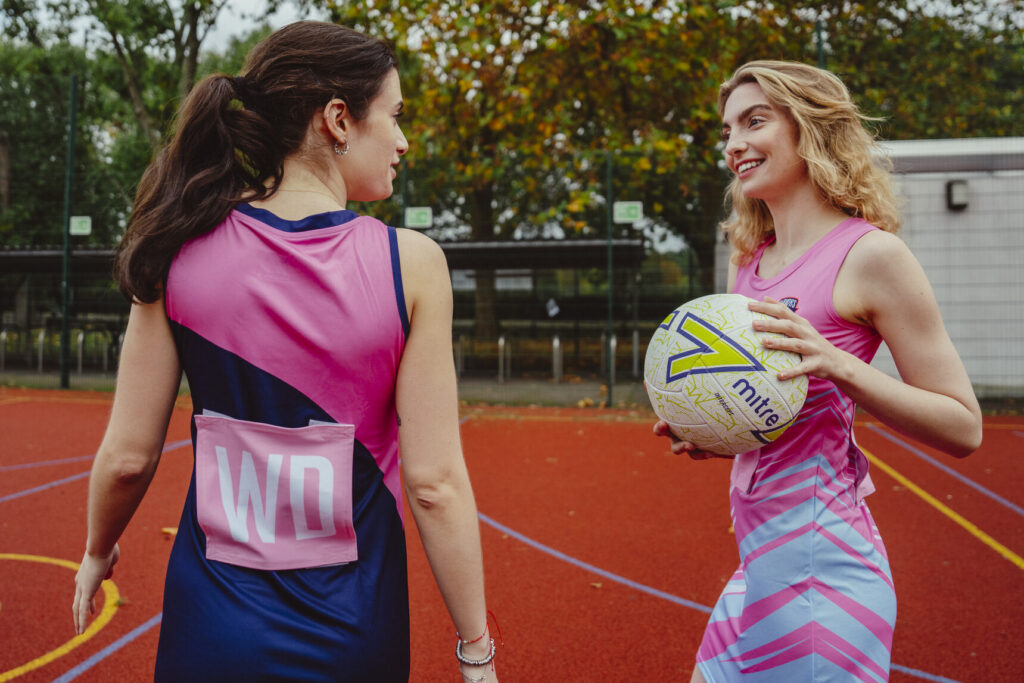 Zwei Frauen in nachhaltiger Sportbekleidung stehen auf einem Netballfeld im Freien. Eine hält einen Netball in der Hand und lächelt die andere an. Im Hintergrund sind Bäume und ein Basketballkorb zu sehen.