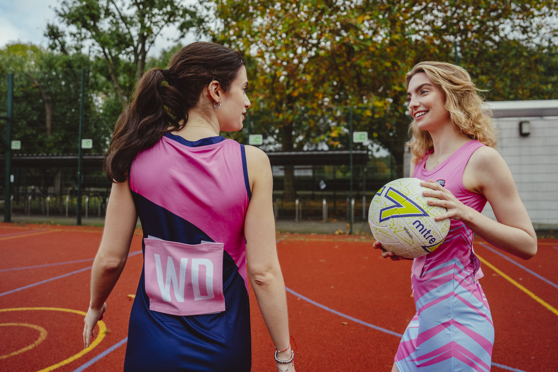 Due donne in divise da netball personalizzate parlano su un campo all'aperto. Una tiene in mano una palla da netball e sorride, mentre l'altra, ripresa di spalle, ha la scritta “WD” sulla schiena. Sullo sfondo si vedono degli alberi e un canestro da basket.