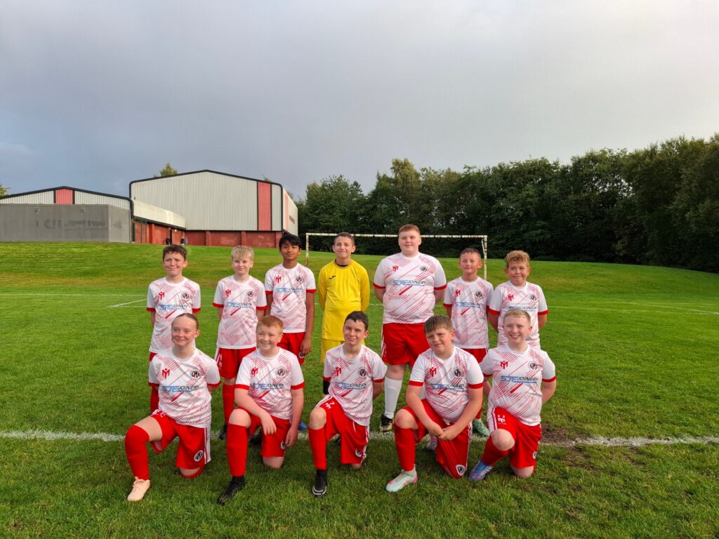 El equipo juvenil del Bellshill Colts posa con una equipación personalizada a juego en blanco y rojo, con el portero vestido de amarillo, en un campo de césped.