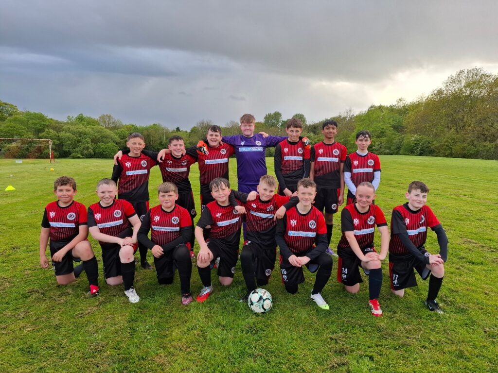 La squadra giovanile di calcio - i Bellshill Colts - posa per una foto di gruppo su un campo in erba, indossando le divise "My Club" in rosso e nero realizzate con tecnica di sublimazione, con i numeri di maglia e i loghi degli sponsor