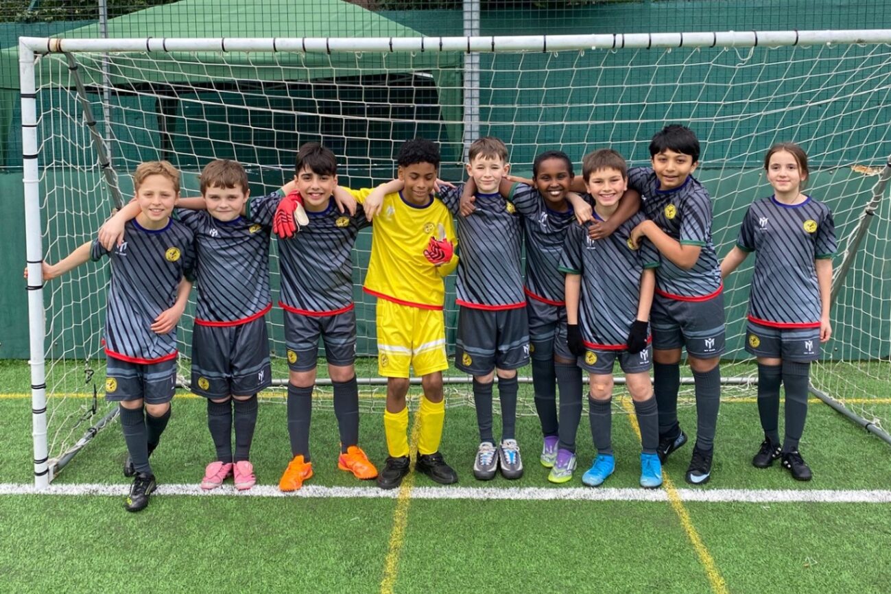 A junior grassroots football team in matching grey and red kits posing together in front of a goal, arms around each other