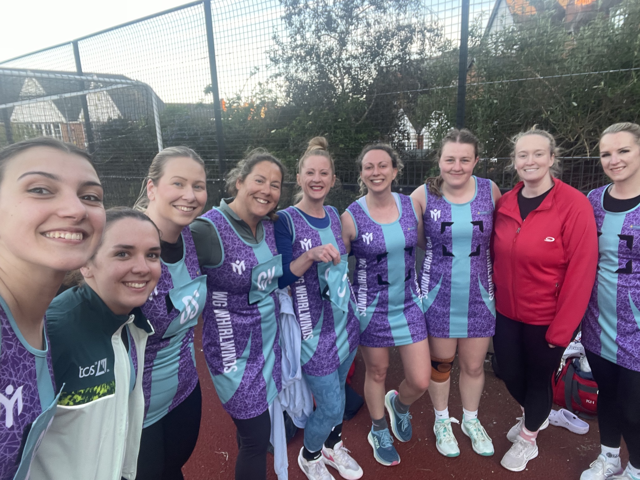 Die Netballmannschaft „WG Whirlwinds“ posiert gemeinsam auf einem Außenplatz in einheitlichen lila- und türkisfarbenen „My Club“-Trikots