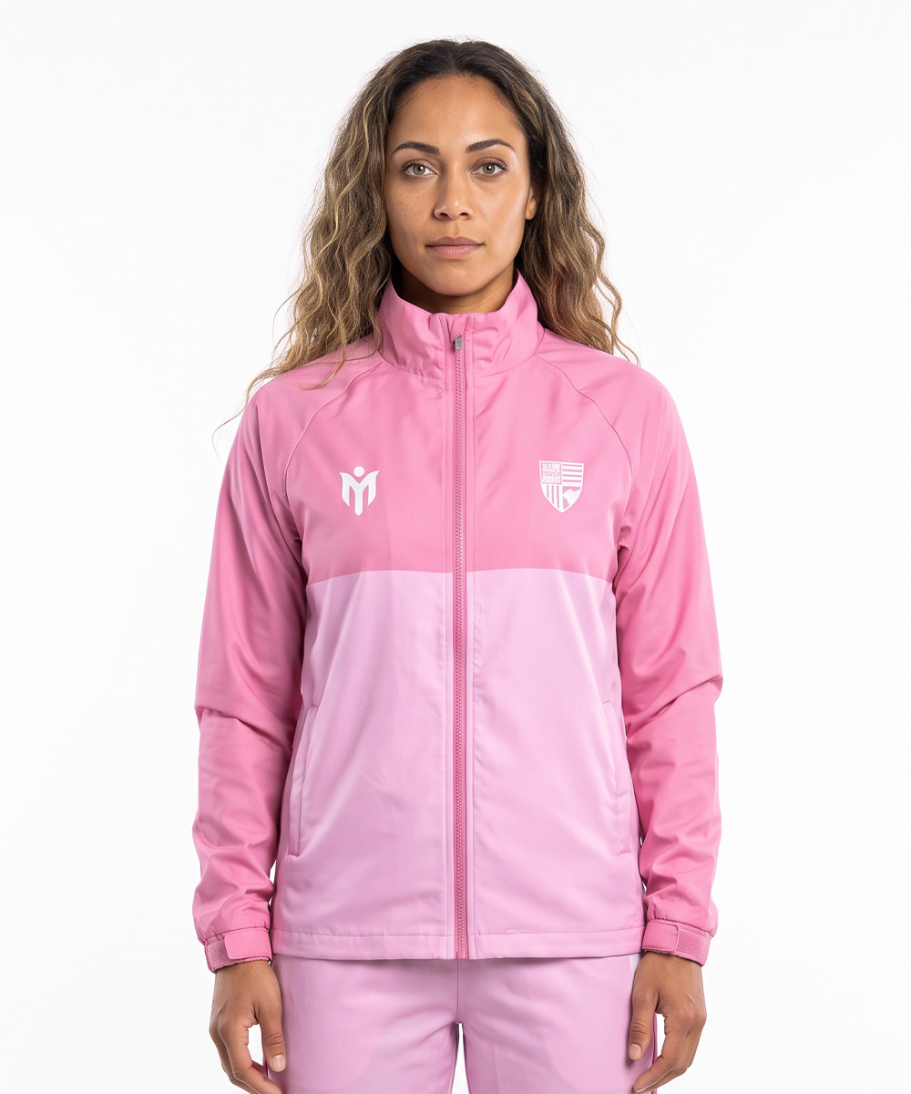 Eine Frau mit langen, welligen Haaren trägt eine rosa Sportjacke mit Reißverschluss und eine dazu passende Hose, die an maßgeschneiderte Softshell-Jacken erinnern. Sie steht vor einem schlichten weißen Hintergrund, blickt nach vorne und hat einen neutralen Gesichtsausdruck.