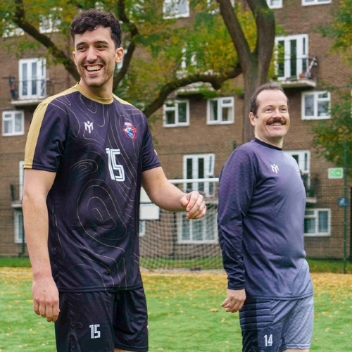 Due uomini in divisa da calcio sono in piedi e sorridono su un campo erboso, con un complesso residenziale e degli alberi sullo sfondo. Uno indossa una maglia nera con il numero 15, mentre l’altro indossa una maglia grigia con il numero 14.