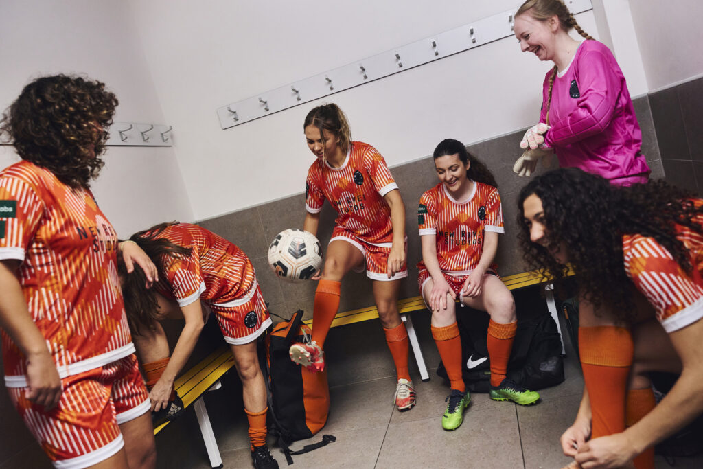 Women's football squad in matching orange custom kits and a pink goalkeeper shirt getting ready in a changing room before a match
