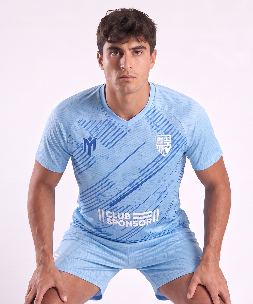 Un uomo con una divisa da calcio azzurra è seduto su una panchina e guarda davanti a sé con espressione seria. La maglia, una delle divise personalizzate della squadra, presenta motivi bianchi e azzurri, uno stemma e il nome dello sponsor del club.