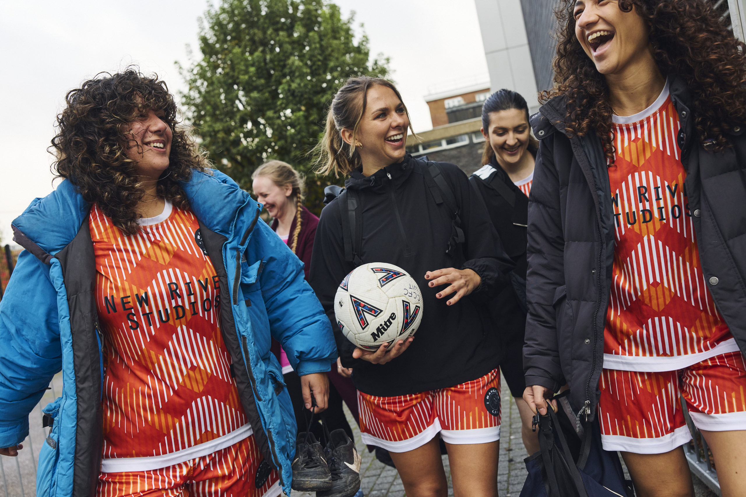 A group of smiling football players wearing orange and white graphic shirts and shorts, one holding a Mitre football.
