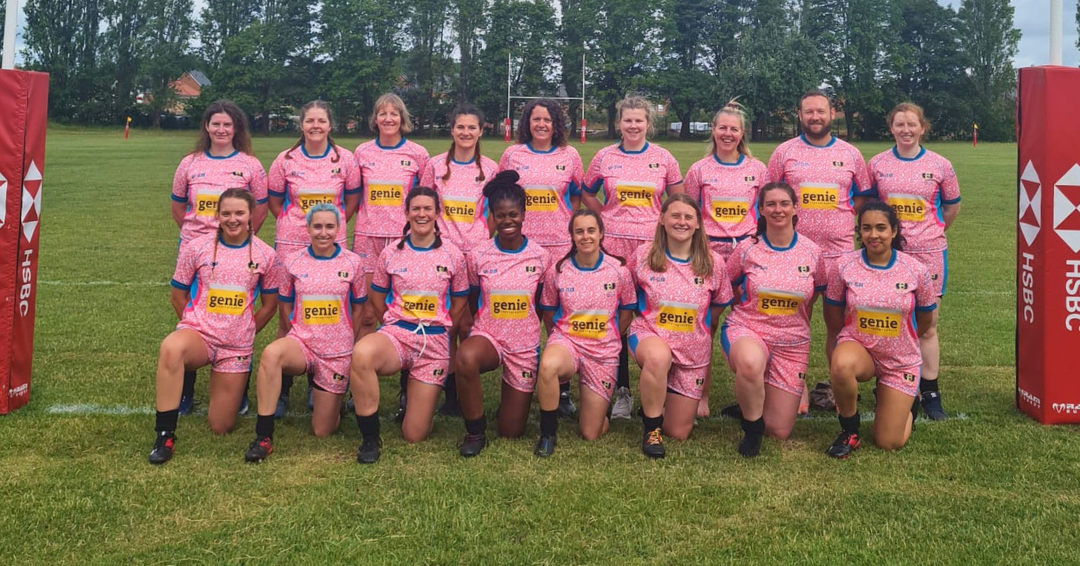 A sports team posing for a group photo on a grass pitch, wearing mud-stained, matching pink patterned rugby kits with the word genie printed on the front.