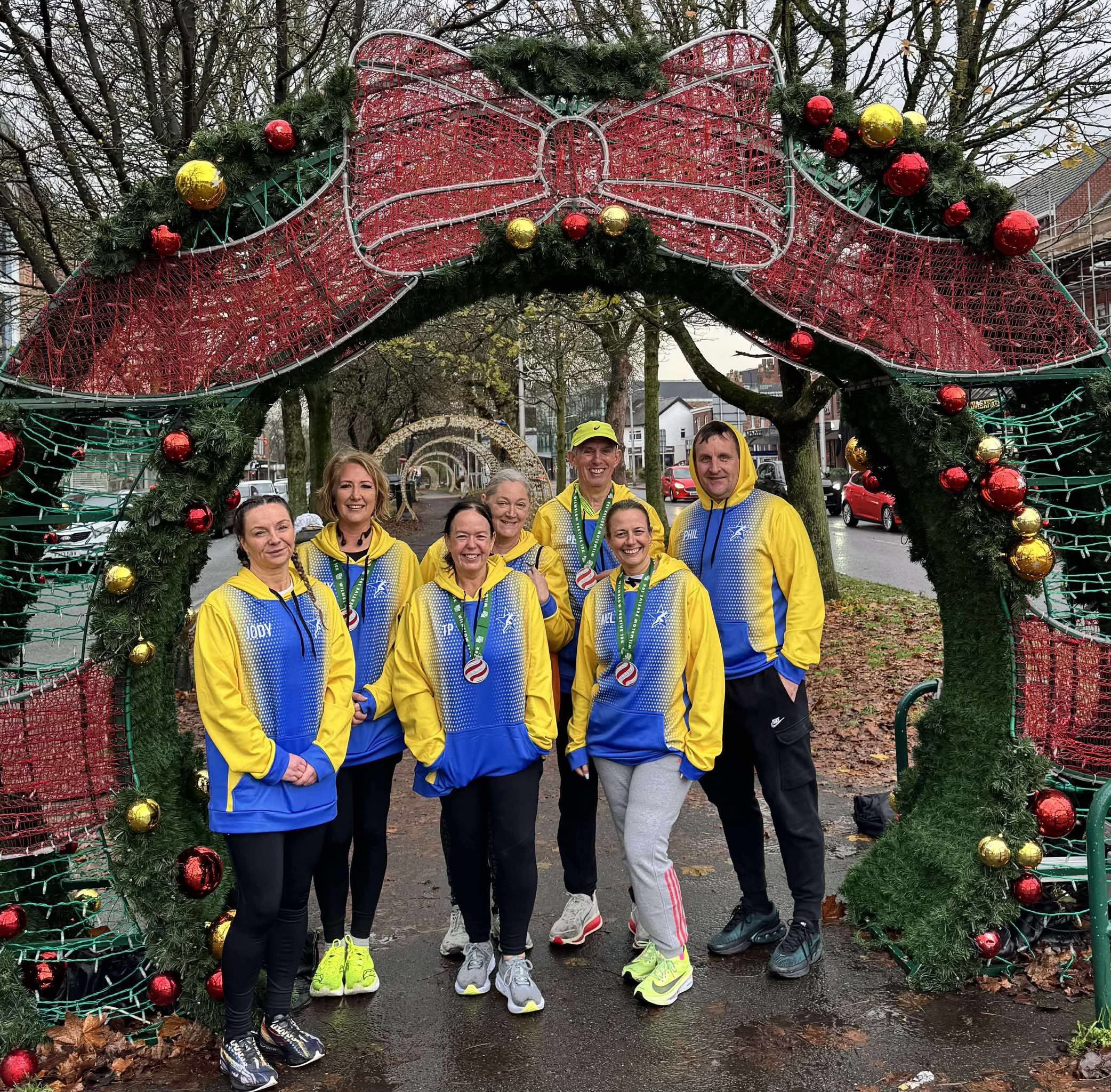 Six people in matching yellow and blue hoodies wear race medals while posing under a large, arched Christmas decoration.