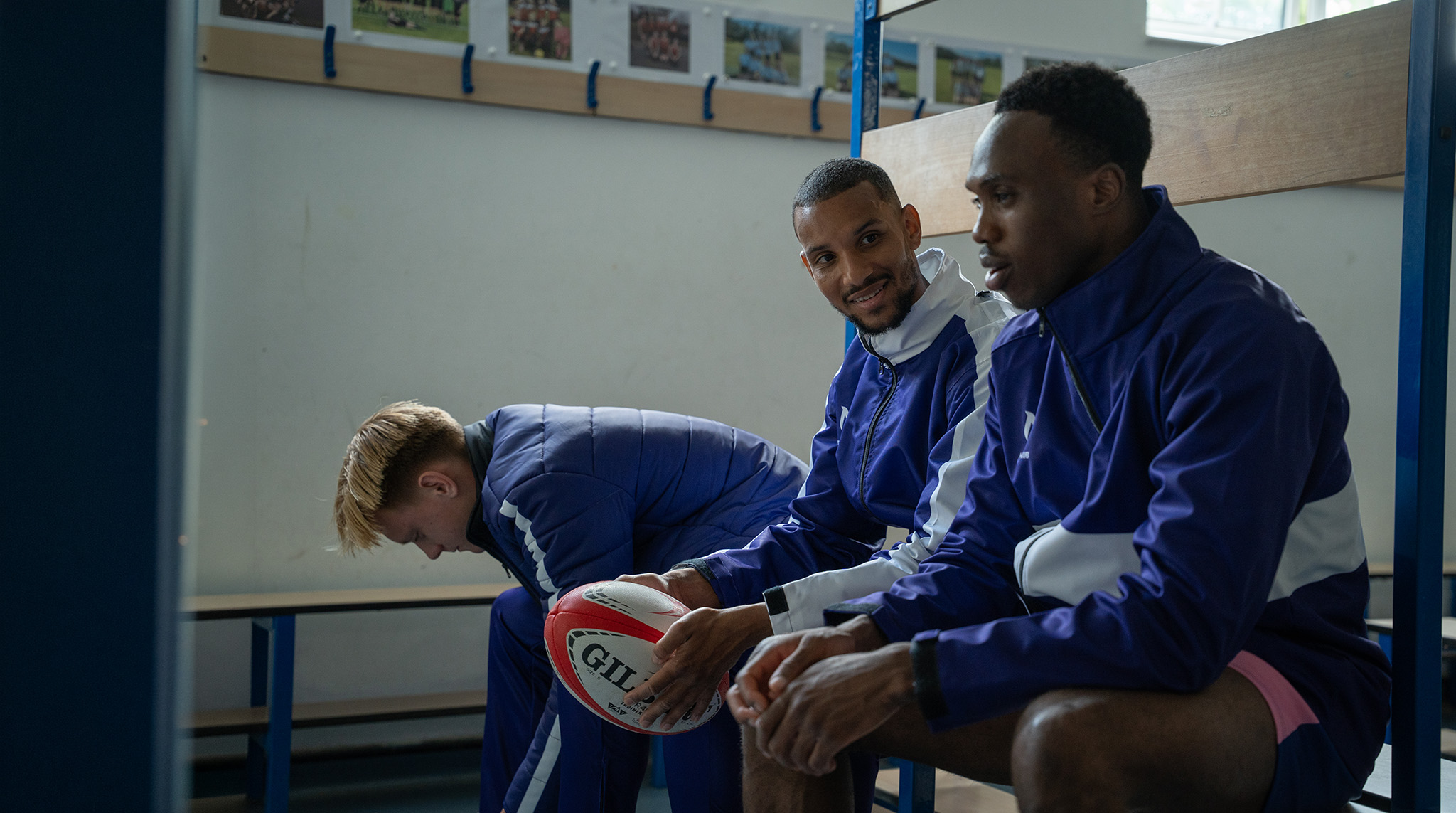 Three rugby players in matching blue team jackets sit on a changing room bench, with one player holding a rugby ball.